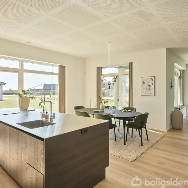 Køkkenø med vask står centralt i et lyst åbent rum. En spisebordgruppe med stole ses ved vinduer, omgivet af grøn have. Ingen tekst til stede.