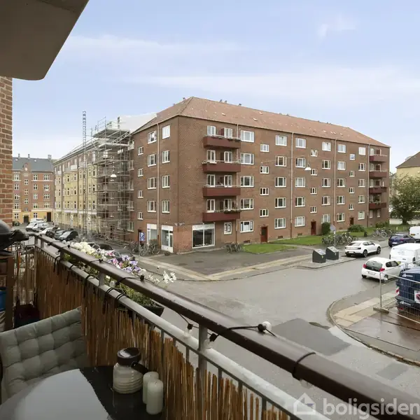 Balkon med grill og møbler står mod rødstensbygninger. På gaden står biler parkeret, og der er byggeri med stilladser på en bygning.