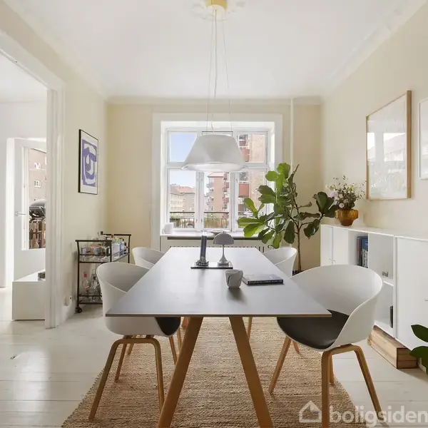 Et spisebord med to moderne stole står på et tæppe. På bordet ligger bøger og en lampe hænger over. Rummet er lyst og indrettet med grønne planter og billeder på væggene.