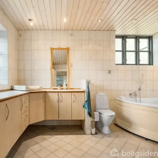 Badeværelse med lyse fliser og træloft. Der er et hjørnebadekar til højre, et toilet ved siden af, og en dobbeltvask med spejl til venstre.