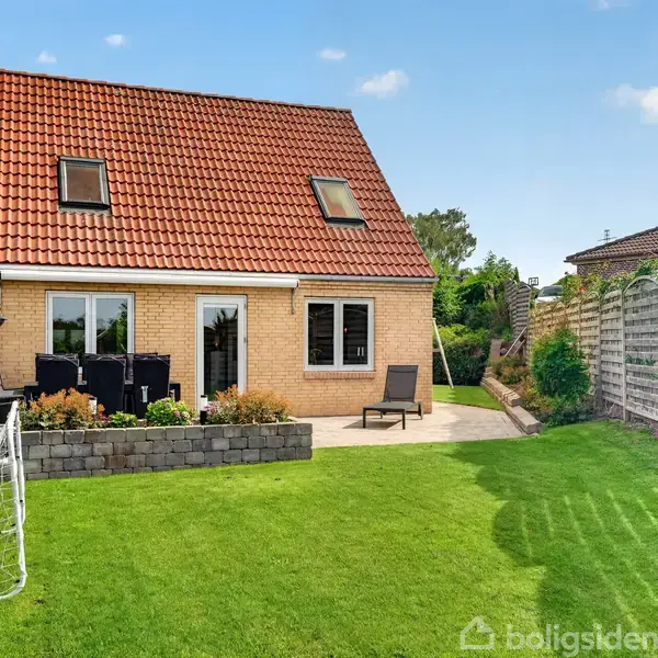 Hus med rødt tegltag og gule murstensvægge. Terrasse med havemøbler foran huset. Græsplæne med fodboldmål i haveomgivelse, omkranset af hække og hegn under klar blå himmel.