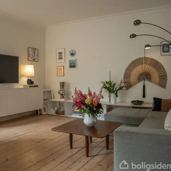 En grå sofa står i en stue med trægulv. Sofaen har puder, og foran er et sofabord med en blomsterbuket. En TV står til venstre, omgivet af lamper og kunst på væggene.