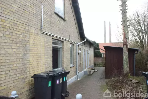 En række skraldespande står på en flisebelagt gang ved siden af en murstensbygning; i nærheden er et lille skur og nogle træer. Baggrunden viser to skorstene.