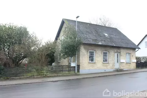 Et murstenshus med skråt tag står ved siden af en vej. Huset har vinduer med hvide rammer og en træport. Omkring er der en lav hegn og tætte buske.