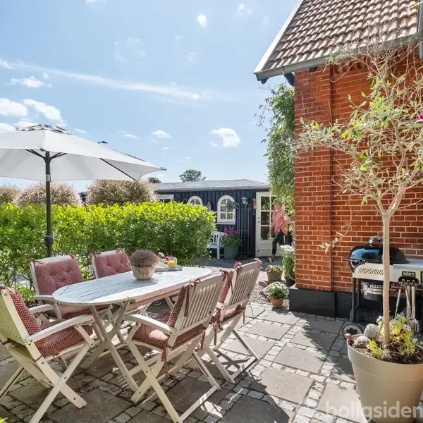 Et bord med stole står under en parasol på en stenbelagt terrasse. Baggrunden viser en rød murstensbygning og grønne planter, hvilket skaber en hyggelig udendørs atmosfære.