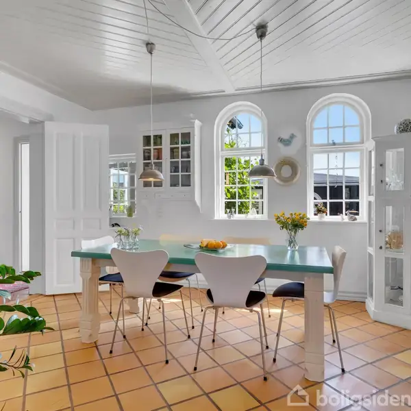 Spisebord med moderne stole står i et lyst rum med terracotta-fliser. To pendellamper hænger over bordet. Store vinduer og et rundt vitrineskab fyldt med dekorationer præger væggen.