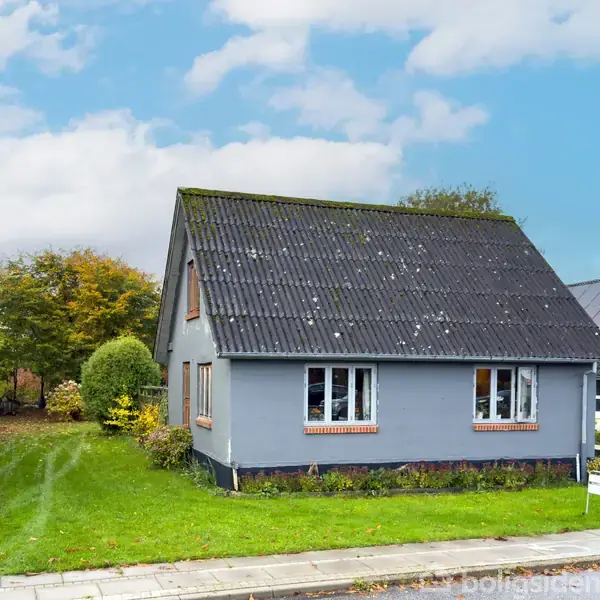 Et gråt hus med et mørkt tag står stille ved siden af en sti. Baggrunden består af træer og en have.