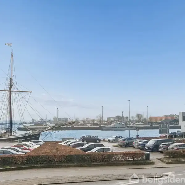 Et skib ligger til kaj, omkranset af parkerede biler på en plads ved havnemiljøet. Bygninger og en vej ses i baggrunden under en klar blå himmel.