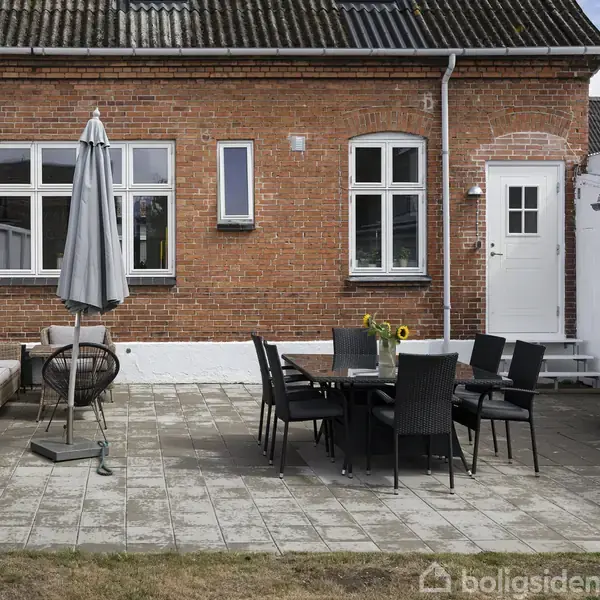 Terrasse med mørkt havebord og stole samt en parasol. Rundt om er der en murstensbygning med hvide vinduer og dør. Der ses en grill og en græsplæne.