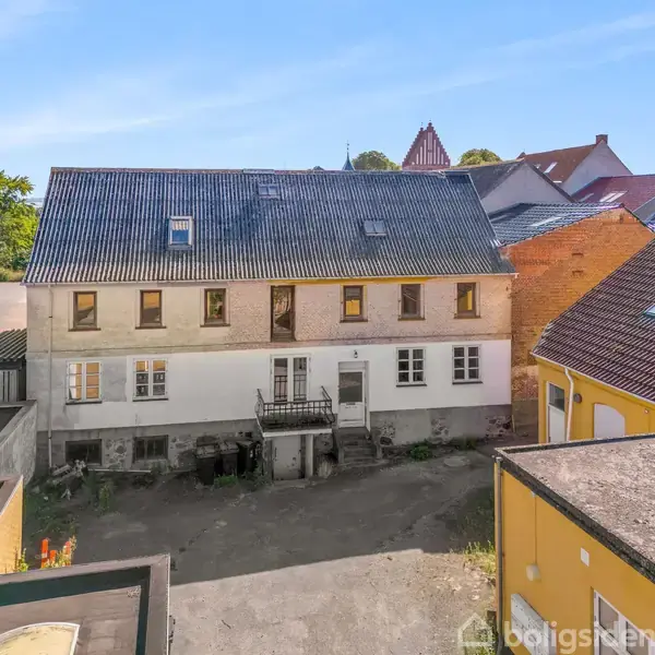 To-etagers hvid bygning med skråt, mørkt tag står i en gård omgivet af flere farverige bygninger under en blå himmel. Der ses træer i baggrunden.