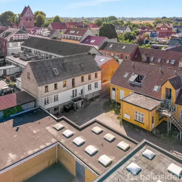 Boligblokke med skråtage står side om side i en by. En hvid bygning og en gul bygning med udvendig trappe ses forrest, omgivet af andre bygninger.