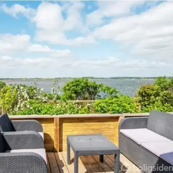 Terrasse med grå havemøbler, herunder to stole og en sofa omkring et bord. Møblerne er placeret, så de tilbyder udsigt over en sø eller fjord med grønne buske og træer i baggrunden.