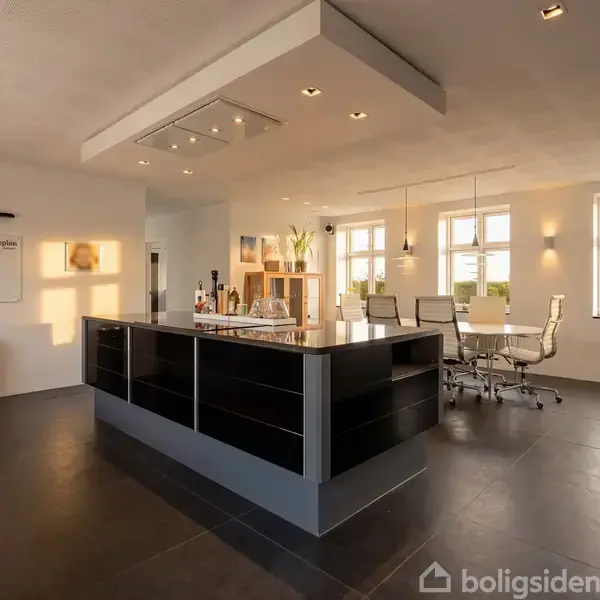 Et moderne køkken har en stor kogeø med sort og hvidt design. Rummets lyse indretning inkluderer spiseplads med fire stole. Tekst på væg: “Fremtiden bygger ifølge engelske synonymer.”