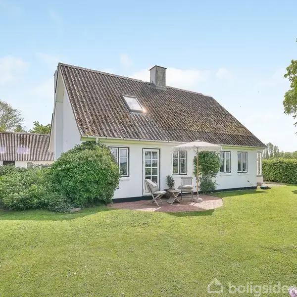 Hus med skrånende tag står stille i en velplejet have med grønne buske og træer. Et lille terrasseområde med bord og stole er synligt foran huset med blå himmel bagved.