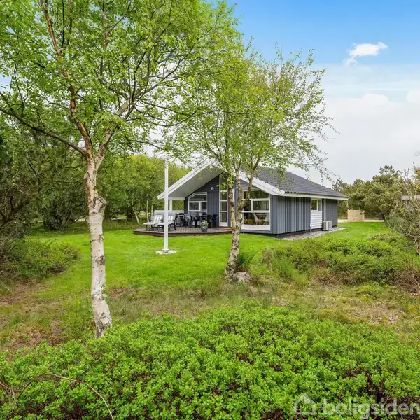 Et blågråt sommerhus står i en grøn have omgivet af høje træer. En terrasse med havemøbler ses foran huset. Klart himmel danner himlen.