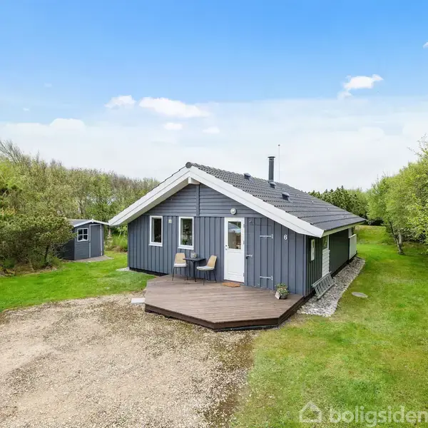Et gråt træhus står på en grøn græsplæne med en lille terrasse foran. På terrassen er to stole, og omgivelserne er fyldt med træer under en klar blå himmel.