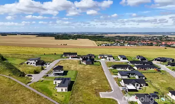 Boligområde med moderne huse ligger langs snoede veje. Området er omgivet af store marker og en by i baggrunden under en blå himmel med spredte skyer.