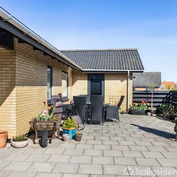 Terrasse med fliser, omgiven af murstensbygning og træhegn. Havemøbler og blomsterkrukker er placeret langs væggen. I hjørnet står en udendørs pejs. Klar blå himmel i baggrunden.