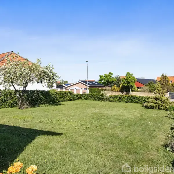 En græsplæne omgivet af hække står stille i en boligområde med røde murstenshuse under en blå himmel. En enkelt lille træ og forskellige buske er plantet langs kanten.