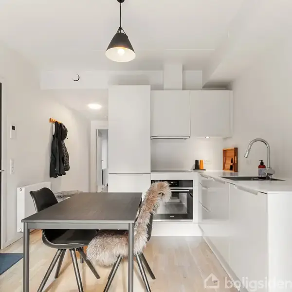 Et moderne køkken-alrum med sorte møbler omkring et spisebord, komfur og vask i en hvid indretning. Belysningen hænger fra loftet, og overtøj hænger ved døren.