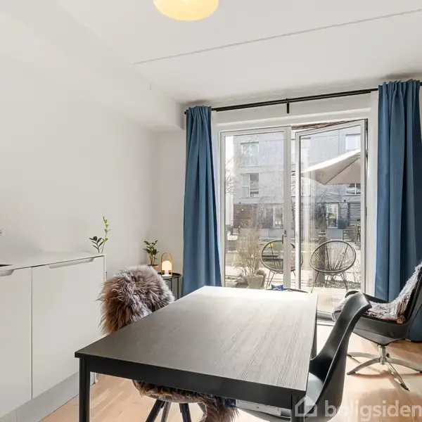 Et sort bord står foran åbne altandøre i et lyst rum med hvide vægge, blå gardiner og køkkenvask til venstre. To stole er ved bordet, planter i hjørnerne.