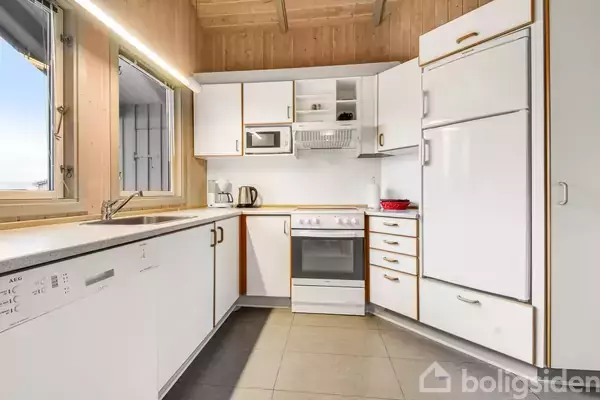 Et hvidt køkken har skabe og skuffer, en ovn, mikrobølgeovn og køleskab, mens solen lyser ind gennem et vindue. Tæppebelagt gulv skaber en skarp og ren atmosfære.