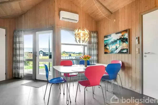 Et spisebord står i et lyst rum, omgivet af farverige stole og en moderne lampe hængende over bordet. Store vinduer viser en udsigt til naturen udenfor.