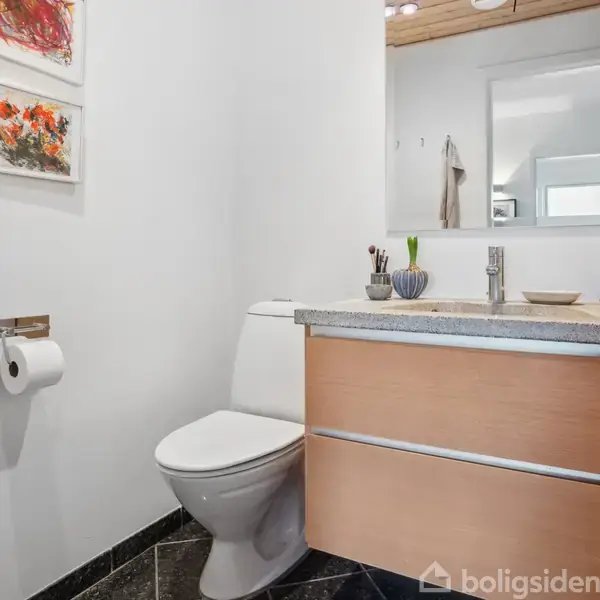 Toilet og vask i et moderne badeværelse. Toilettet står ved siden af en træskabssektion med en vask ovenpå. Der er et stort spejl og kunst på væggen.