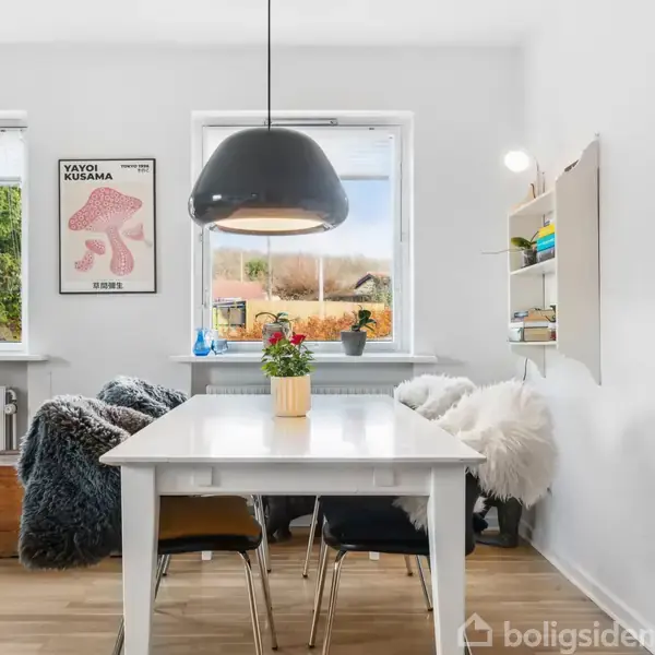 Et hvidt spisebord med sorte stole står i et lyst rum. En sort pendellampe hænger over bordet. To vinduer viser udsigt udenfor. Til venstre hænger en plakat med teksten "Yayoi Kusama".