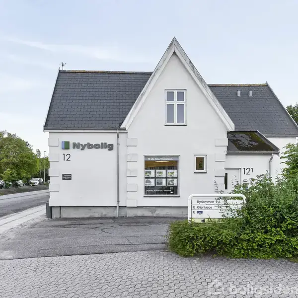 En hvid bygning med skiltet "Nybolig," viser et displayvindue med ejendomslister. Bygningen ligger ved en gade omgivet af grønne buske og træer, med adressenummeret "12."
