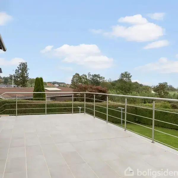 En rummelig, flisebelagt terrasse med metalrækværk strækker sig fra et hus. Terrassen har en vid udsigt over en grøn have og et bakket landskab i det fjerne.