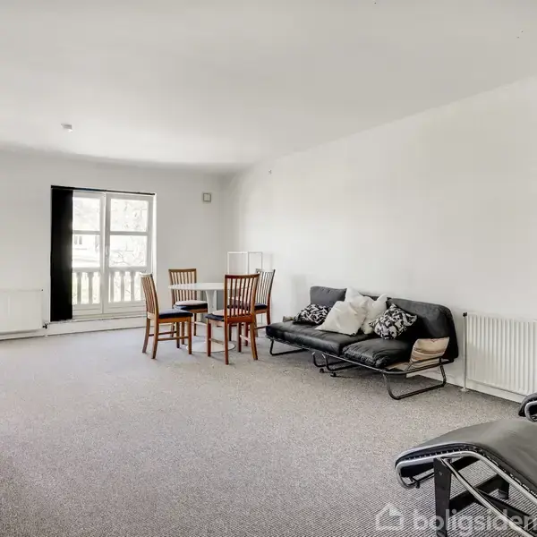 En stue med en sofa og puder langs en hvid væg. To stole og et bord står ved et vindue. En skænk og lænestole er også til stede. Ingen tekst fundet.