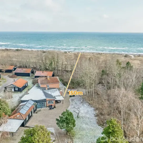 Hus tæt på strand, markeret med linje på 150 m afstand, omgivet af træer og flere huse. Hav i baggrunden, skaber en naturskøn kystlandskab.