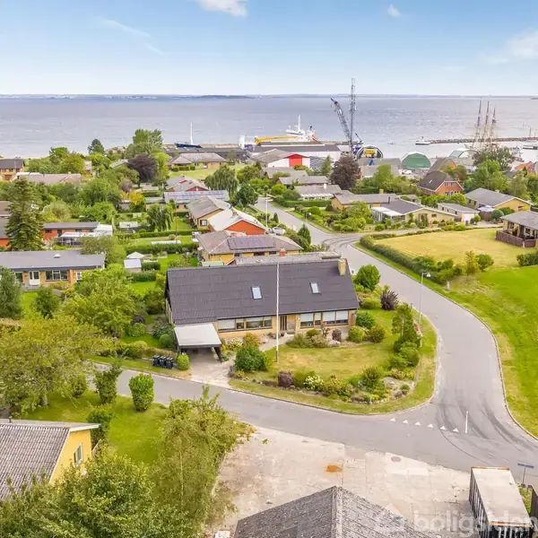 Et boligområde med huse omgivet af grønne haver og træer. En vej snoer sig gennem nabolaget. I baggrunden ses en havn med skibe og en udsigt over havet.