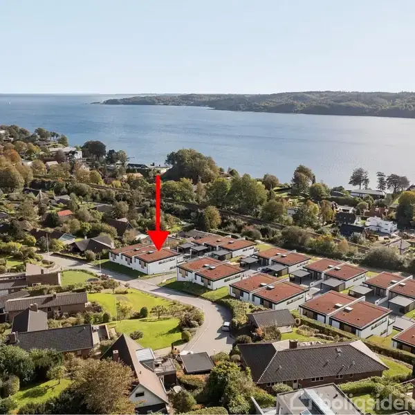 En rød pil peger ned mod et hus i et boligområde. Området er fyldt med ensartede huse med røde tage og ligger tæt ved en kyst med skovklædte områder.