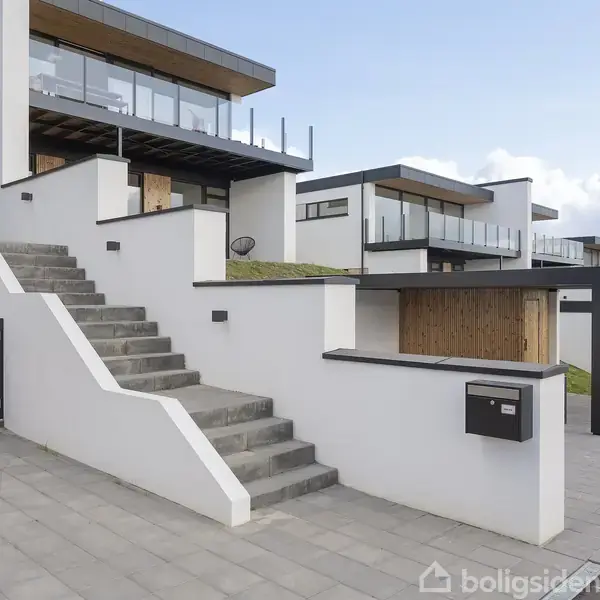 Moderne hvid bygning med store vinduer og terrasser. Trapper fører op til indgangen. Gårdspladsen har flisebelægning, og baggrunden viser flere lignende bygninger. Postkasse monteret på væggen.