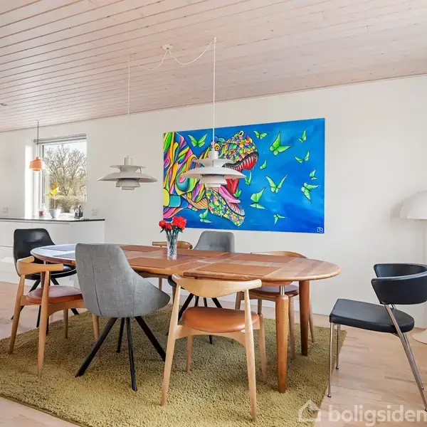 Et ovalt træbord med stole står på et tæppe midt i et lyst spisekøkken. En farverig tigerplakat hænger på væggen. Vinduet viser en have udenfor.
