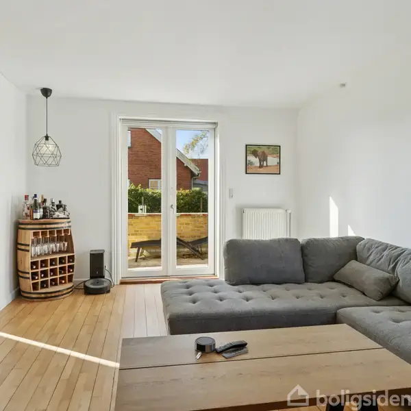 Stor, grå hjørnesofa står i en lys stue med trægulv. En træbord med en fjernbetjening er foran sofaen. Dør fører til terrasse med bord og stole udenfor. Barområde til venstre.