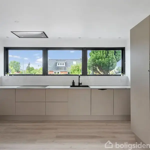 Moderne køkken med beige skabe og sort vandhane, set foran et stort vindue. Udenfor ses et grønt træ og et hus. Højre side har en indbygget ovn.