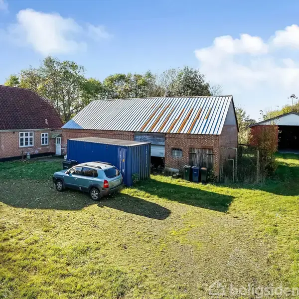 En bil holder parkeret foran en stor lade med bliktag. Til venstre står et rødt murstenshus. Hele scenen er omgivet af træer, græs og en blå container.