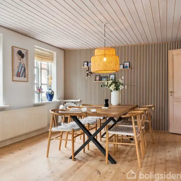 Et spisebord med stole står i et lyst spiseområde. En gul lampe hænger over bordet. Der er gulvlange vinduer, en stribet væg og dekorationer på reolen. En plakat hænger på væggen.