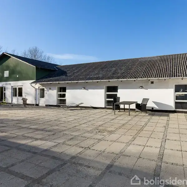 En hvidmalet bygning med sort tag står stille på en stor, flisebelagt gårdsplads. Bord og stole ses foran bygningen, omgivet af en klar blå himmel og træer.