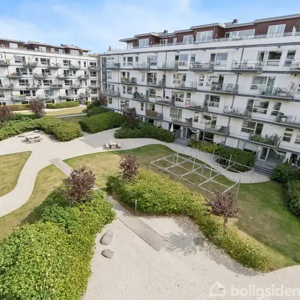 Lejlighedskompleks omgiver en indre gårdhave, hvor grusstier, grønne buske og picnicborde i sollys skaber et indbydende miljø. Balkoner og store vinduer præger bygningens facade.