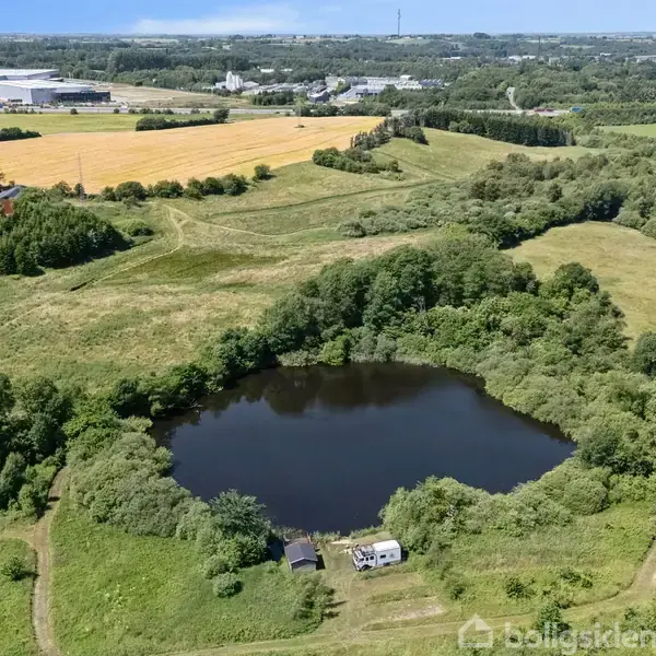 Sø omgivet af grønt landskab med stier og træer. To små bygninger ses nær søen. I baggrunden marker og en industriel bygning.