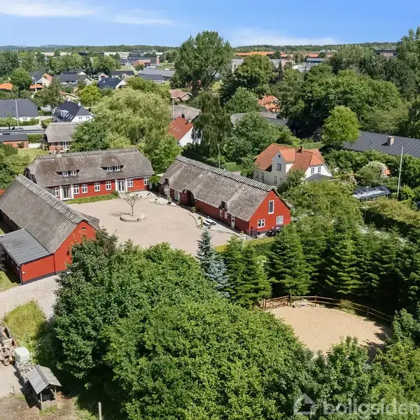 Rødt bondehus med stråtag står i midten omgivet af frodig beplantning. Flere røde bygninger ligger rundt omkring på gårdspladsen, der er belagt med sten og omgivet af grønne træer.