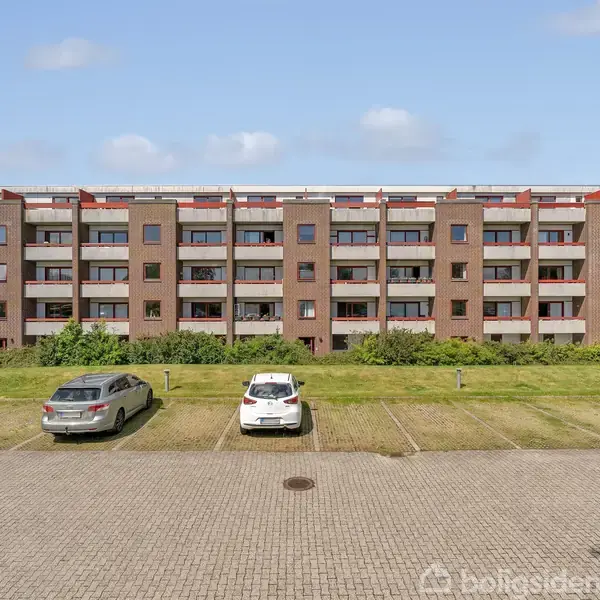 Lejlighedskompleks står stille ved siden af en parkeringsplads med flere biler. Omgivet af træer og buske under en klar blå himmel.
