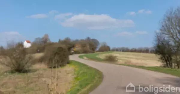Snoet landevej strækker sig gennem åbne, grønne marker under en blå himmel med få skyer. En hvid bygning med rødt tag ses til venstre i baggrunden.