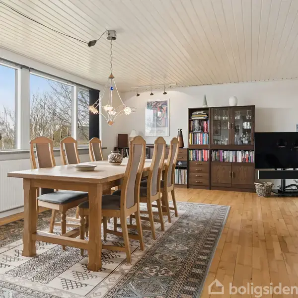 Stort spisebord med træstole står i en lys stue. Bordet er dækket med en lys dug, og omgivet af store vinduer. Til højre er en reol med bøger.