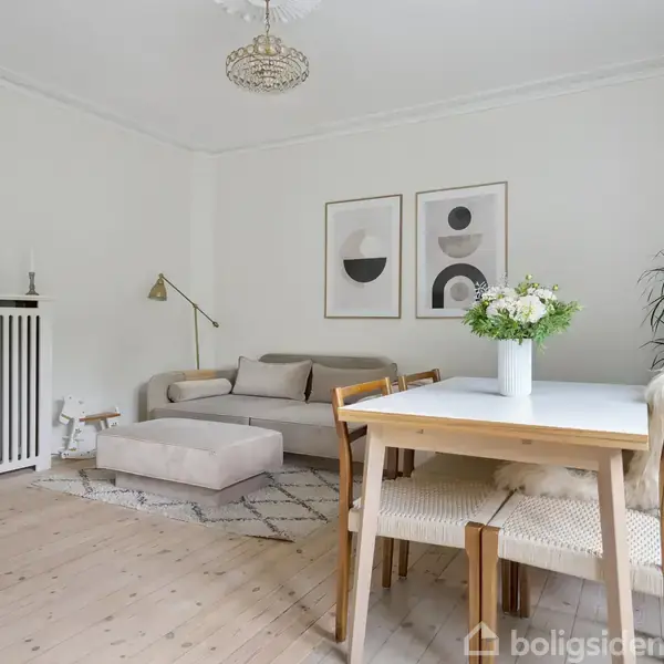 Sofa står i lys stue ved siden af radiator og moderne billeder. Rundt sofabord med lampe ved siden af, lille reol på væggen og planter nær spisebord med blomster.