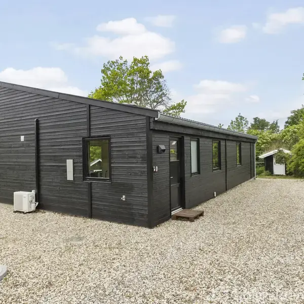 Et sort træhus står stille omgivet af grønne buske og træer på en grusvej. Huset har et skråtag og flere vinduer langs siden.
